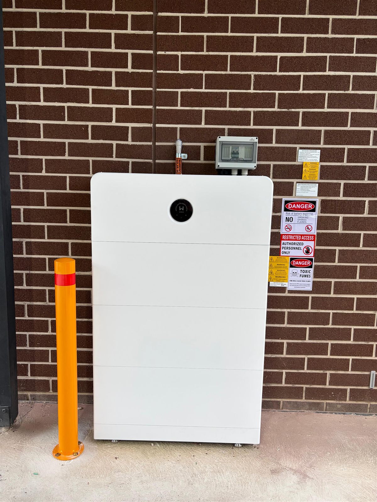Home battery system installed on an exterior brick wall with safety bollard and signage